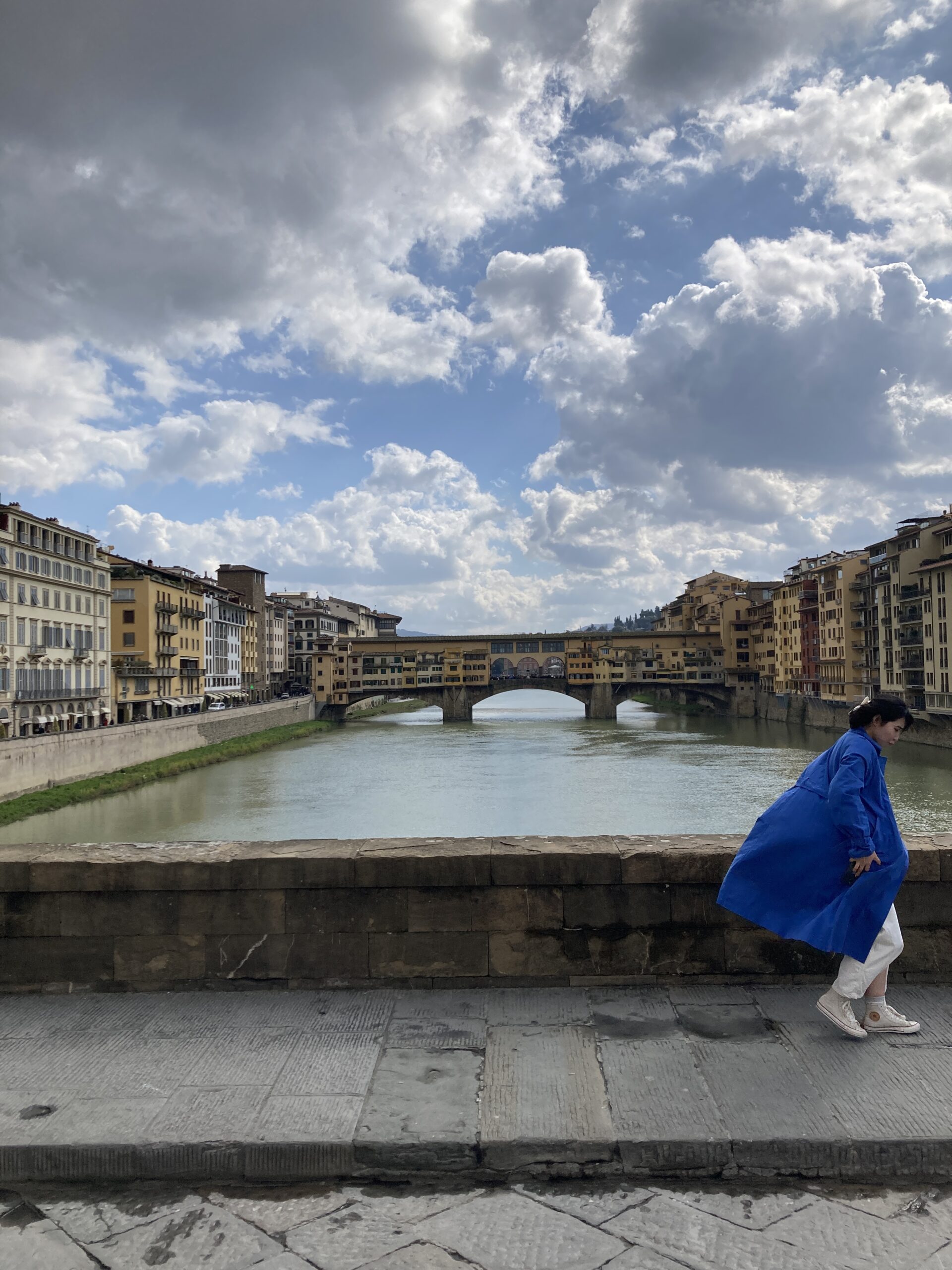 A view of a bridge across a river, with a cloudy sky above. A woman is seen in the foreground, her blue coat billowing behind her.